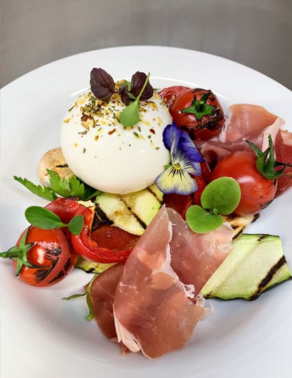 Burrata-Käse mit gegrillten Tomaten, Zucchinischeiben, Parmaschinken und essbaren Blüten auf weißem Teller