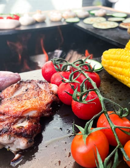 Gegrilltes Hähnchen, Tomaten am Strunk und Maiskolben auf Grill mit Flammen im Hintergrund