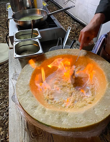 Flambiertes Gericht in einem ausgehöhlten Parmesanrad, daneben Kochutensilien und Soßenbehälter