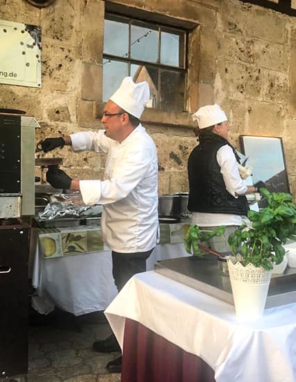 Zwei Köche in weißen Uniformen und Kochmützen bereiten Pizza an einem mobilen Pizzaofen vor, im Vordergrund ein Tisch mit weißer Tischdecke und Basilikumtopf